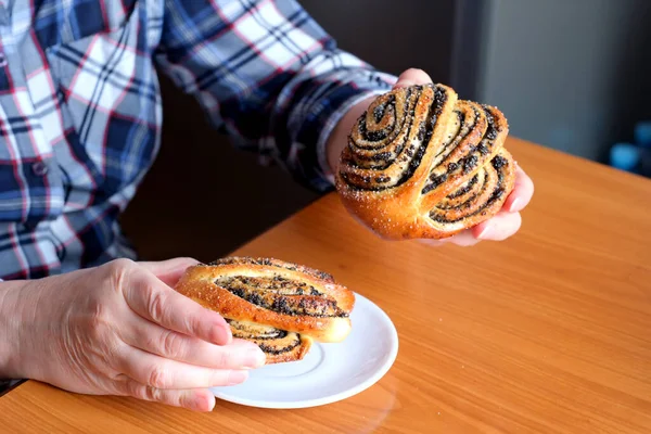 The hands of an elderly woman operate a sweet bun with poppy seeds in the kitchen at a brown table, without a face, close-up