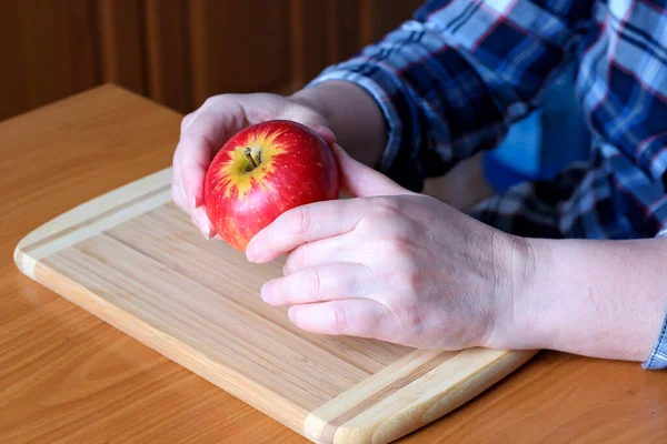 The hands of an elderly woman hold a red ripe tasty juicy apple in the kitchen at a brown table, without a face, close-up