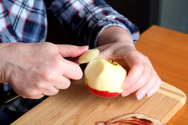 The hands of an elderly woman peel a red ripe tasty juicy apple with a knife in the kitchen at a brown table, without a face, close-up