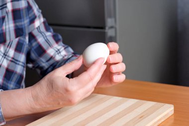 Yaşlı bir kadının elleri kaynamış bir yumurta gösteriyor ve kahverengi bir masadaki mutfakta soyuyorlar, yüzü yok, yakın plan.