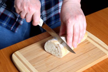 Yaşlı bir kadının elleri, kahverengi bir masanın mutfağında yüzsüz bir bıçakla beyaz buğday ekmeği kesiyor.