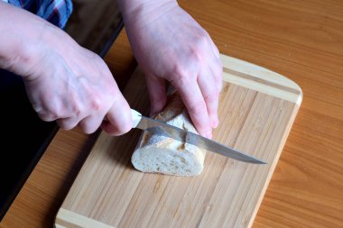 Yaşlı bir kadının elleri, kahverengi bir masanın mutfağında yüzsüz bir bıçakla beyaz buğday ekmeği kesiyor.