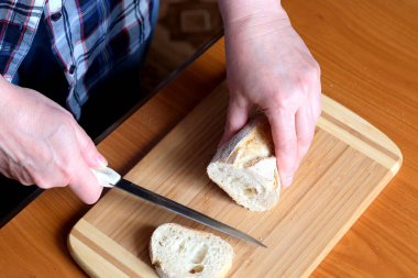 Yaşlı bir kadının elleri, kahverengi bir masanın mutfağında yüzsüz bir bıçakla beyaz buğday ekmeği kesiyor.