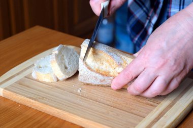 Yaşlı bir kadının elleri, kahverengi bir masanın mutfağında yüzsüz bir bıçakla beyaz buğday ekmeği kesiyor.