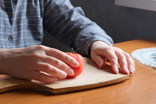 An elderly woman\'s hands hold a red ripe tomato in the kitchen at a brown table, no face, close-up