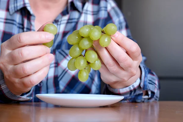 The hands of an elderly woman holds a small brush of white seedless grapes and displays them in the kitchen at a brown table, without a face, close-up