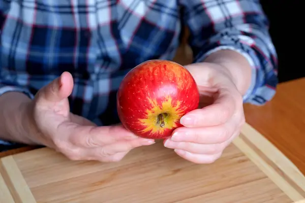 The hands of an elderly woman hold a red ripe tasty juicy apple in the kitchen at a brown table, without a face, close-up