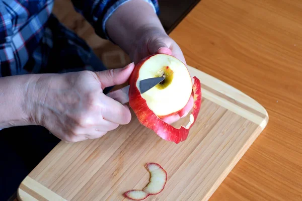 The hands of an elderly woman peel a red ripe tasty juicy apple with a knife in the kitchen at a brown table, without a face, close-up