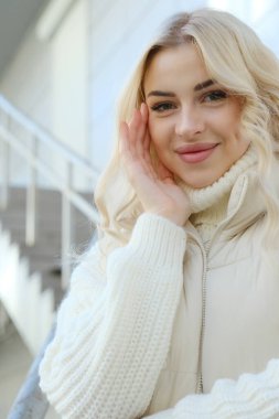 Şehirde güneşli bir sonbahar yürüyüşünde beyaz bir kazak ve ceket içinde uzun saçlı güzel bir sarışının portresi.