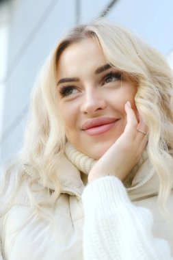 Şehirde güneşli bir sonbahar yürüyüşünde beyaz bir kazak ve ceket içinde uzun saçlı güzel bir sarışının portresi.