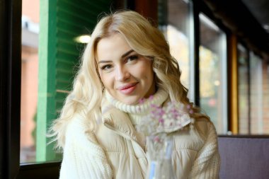 Uzun saçlı, beyaz kazaklı ve ceketli güzel bir sarışının portresi bir kafedeki masada dinleniyor.