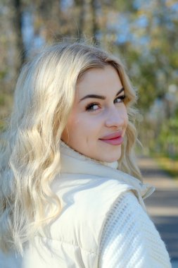 Parkta güneşli bir sonbaharda beyaz bir kazak ve ceket içinde uzun saçlı güzel bir sarışının portresi.