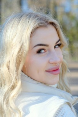 Parkta güneşli bir sonbaharda beyaz bir kazak ve ceket içinde uzun saçlı güzel bir sarışının portresi.