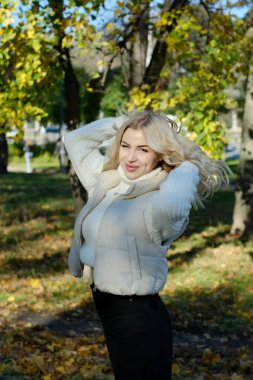 Parkta güneşli bir sonbaharda beyaz bir kazak ve ceket içinde uzun saçlı güzel bir sarışının portresi.