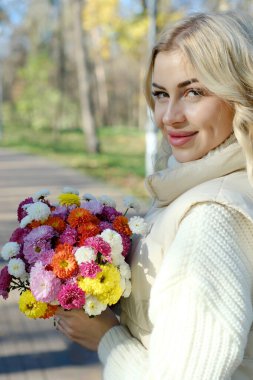 Güneşli bir sonbaharda, bir şehir parkında elinde bir buket çiçekle beyaz kazak ve ceket giyen güzel bir sarışının portresi.