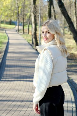 Parkta güneşli bir sonbaharda beyaz bir kazak ve ceket içinde uzun saçlı güzel bir sarışının portresi.