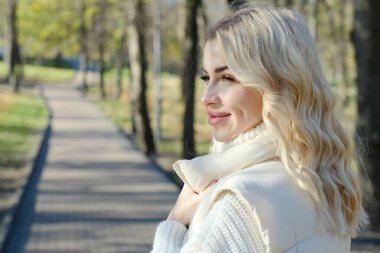 Parkta güneşli bir sonbaharda beyaz bir kazak ve ceket içinde uzun saçlı güzel bir sarışının portresi.