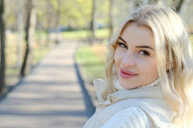 Şehirde güneşli bir sonbahar yürüyüşünde beyaz bir kazak ve ceket içinde uzun saçlı güzel bir sarışının portresi.