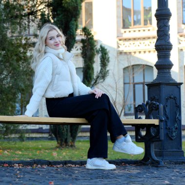 Şehirde güneşli bir sonbahar yürüyüşünde beyaz bir kazak ve ceket içinde uzun saçlı güzel bir sarışının portresi.