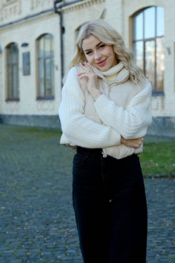 Şehirde güneşli bir sonbahar yürüyüşünde beyaz bir kazak ve ceket içinde uzun saçlı güzel bir sarışının portresi.