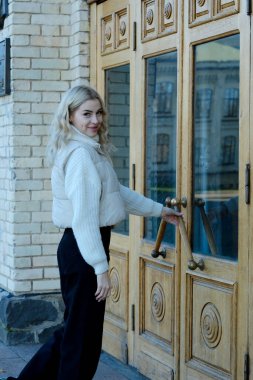 Binanın ön girişinin yanında beyaz kazaklı, uzun saçlı, güzel bir sarışının portresi.