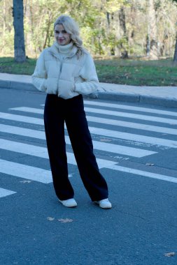 Şehirde güneşli bir sonbahar yürüyüşünde beyaz bir kazak ve ceket içinde uzun saçlı güzel bir sarışının portresi.