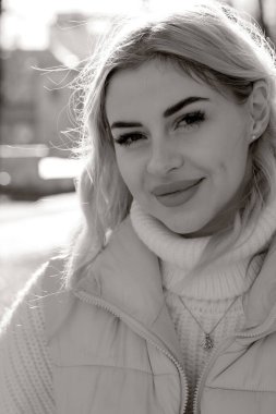 Güneşli bir sonbaharda şehir parkında siyah beyaz bir resimde beyaz kazak ve ceket giyen uzun saçlı güzel bir sarışının portresi.