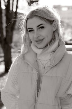 Güneşli bir sonbaharda şehir parkında siyah beyaz bir resimde beyaz kazak ve ceket giyen uzun saçlı güzel bir sarışının portresi.