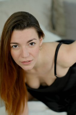 Uzun kızıl saçlı, siyah gömlekli, stüdyoda yatağın üzerinde poz veren genç bir kız.