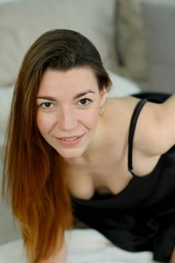 Uzun kızıl saçlı, siyah gömlekli, stüdyoda yatağın üzerinde poz veren genç bir kız.