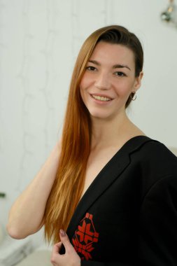 Uzun kızıl saçlı, siyah gömlekli, stüdyoda yatağın üzerinde poz veren genç bir kız.