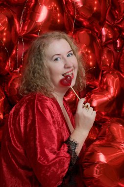 Kırmızı cüppeli, yaşlı, sarışın, kıvırcık bir kadın kalp şeklinde balonlarla stüdyoda poz veriyor..