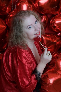 Kırmızı bornozlu, yaşlı, güzel, kıvırcık saçlı bir kadın stüdyoda kalp şeklinde balonlarla poz veriyor..