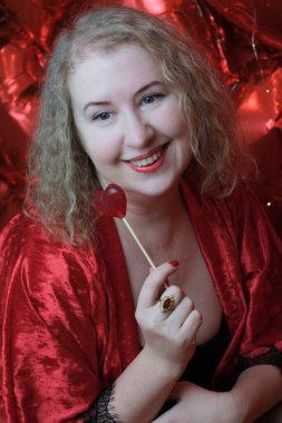 Kırmızı cüppeli, yaşlı, sarışın, kıvırcık bir kadın kalp şeklinde balonlarla stüdyoda poz veriyor..