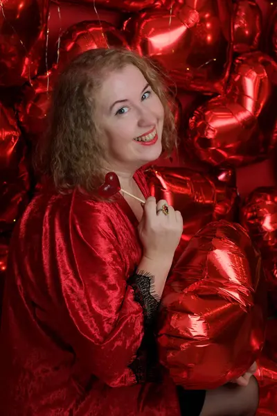 Kırmızı cüppeli, yaşlı, sarışın, kıvırcık bir kadın kalp şeklinde balonlarla stüdyoda poz veriyor..