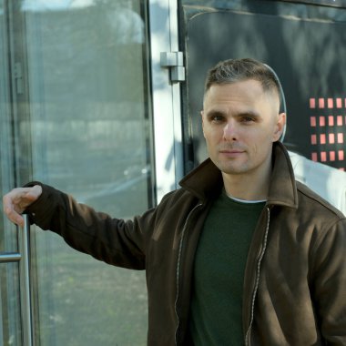 Kahverengi ceketli bir adam, açık havada modern cam bir binanın yanında güven içinde poz veriyor..