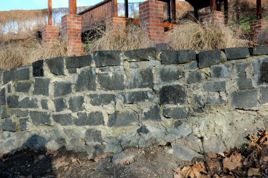 Yıpranmış taşlar ve yakınlardaki uzun otları gösteren taştan bir duvar..