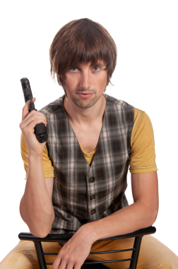 Genç bir adam bir sandalyede oturuyor, kendine güvenerek bir silah tutuyor ve kameraya bakıyor..