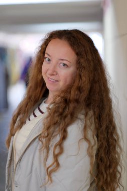 Kıvırcık saçlı bir kadın, hafif ışıklı bir koridorda gülümsüyor ve sıcaklığını yayıyor.