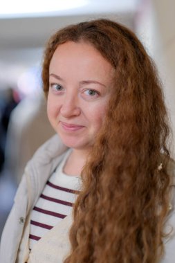 Uzun kıvırcık saçlı, neşeli bir kadın. İçerideyken sıcacık gülümsüyor. Mutluluğu kucaklıyor.