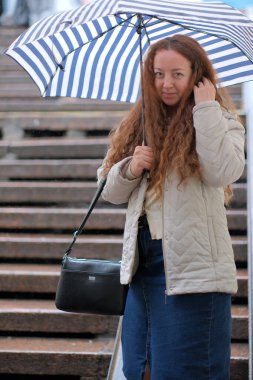 Islak basamaklarda duran bir kadın yağmura karşı çizgili bir şemsiye tutuyor..
