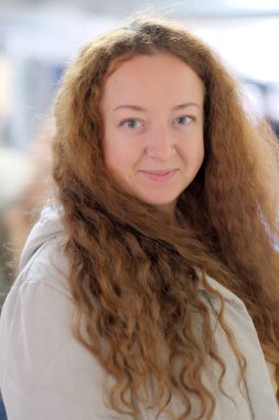 Kıvırcık saçlı genç bir kadın hareketli bir ortamda sıcak, davetkar bir gülümseme sunar.