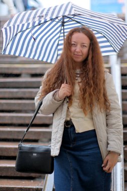 Uzun kıvırcık saçlı neşeli bir kadın elinde çizgili bir şemsiyeyle ıslak basamaklardan aşağı iniyor.