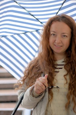 Neşeli genç bir kadın yağmurlu bir günde gülümserken çizgili bir şemsiye tutar.