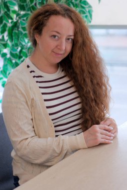 Kıvırcık kahverengi saçlı bir kadın masada oturuyor, rahat bir odada usulca gülümsüyor..