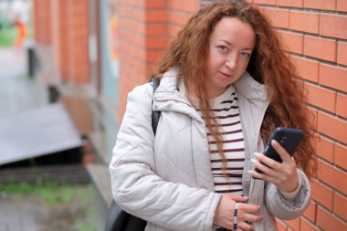 Açık renk ceket giymiş bir kadın duvara yaslanmış telefonuna odaklanmış..