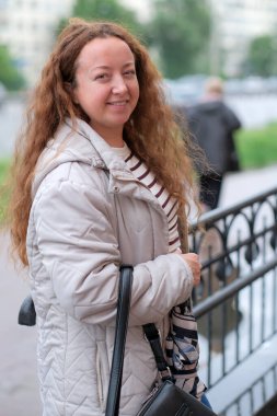 Kıvırcık saçlı bir kadın nehir kenarında yürürken gülümsüyor, doğanın tadını çıkarıyor..