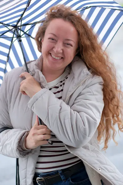 Dalgalı saçlı bir kadın açık havada çizgili bir şemsiye tutarken parlak bir şekilde gülümsüyor..