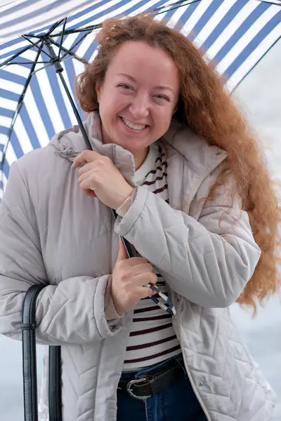 Rahat ceketli neşeli bir kadın çizgili bir şemsiyeyle duruyor, neşeyle gülümsüyor..