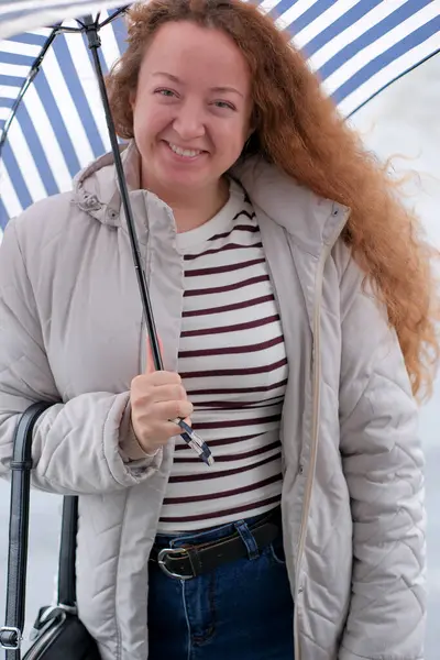 Neşeli bir kadın dışarıda duruyor, çizgili bir şemsiye tutuyor ve yağmura gülümsüyor..